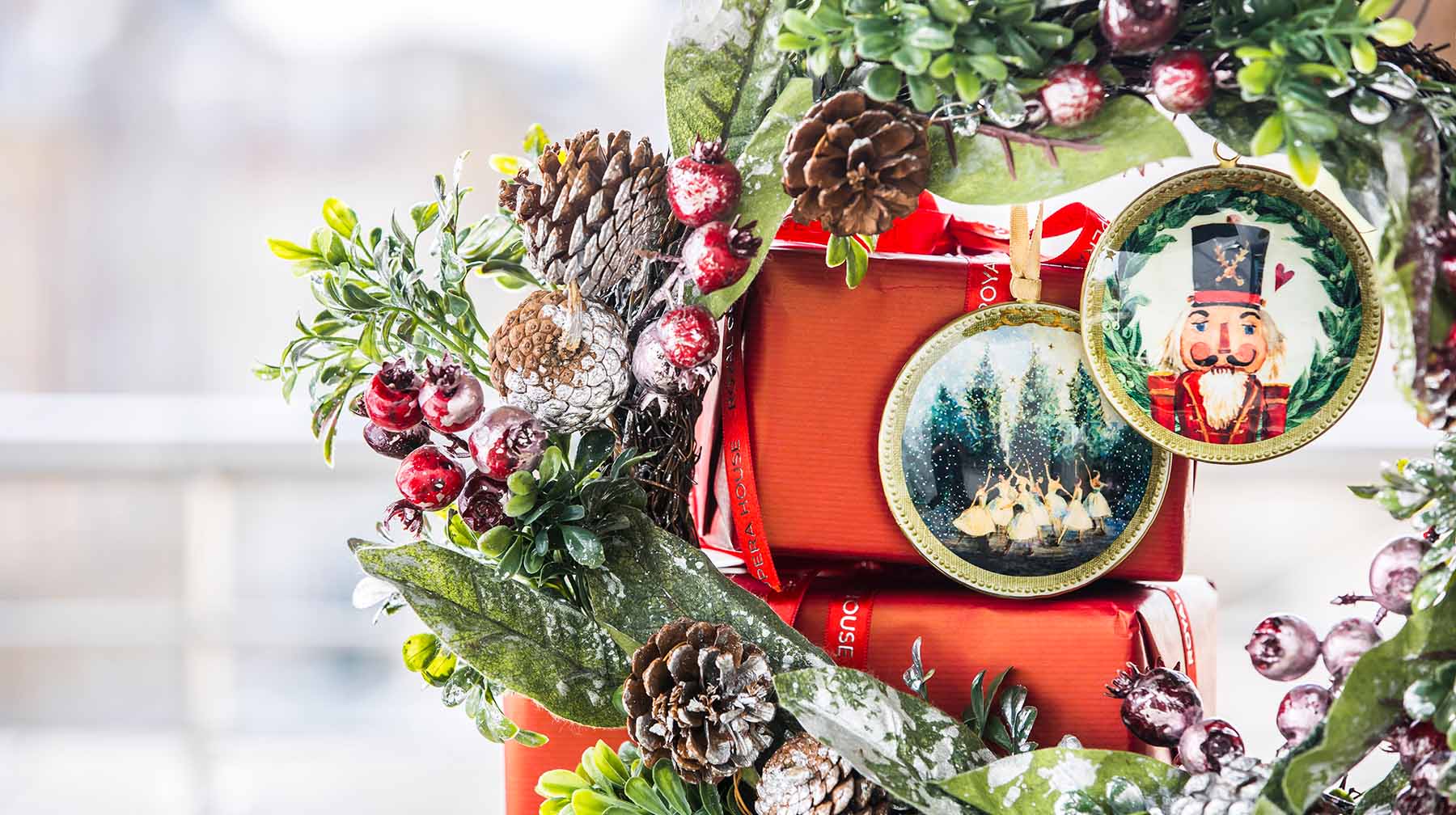 Two Christmas decorations hanging from a wreath in front of a stack of wrapped red gifts.